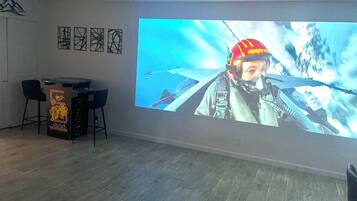 Ein Zimmer mit einem großen Bildschirm, der einen Kampfpiloten zeigt. An der Wand hängen drei gerahmte abstrakte Kunstwerke und ein kleiner Tisch mit Stühlen.