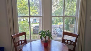 A dining area with a round wooden table and four chairs, a potted plant on the table, and a view of a residential street through large windows. - Duck Pond Getaway (Gainesville)