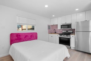 A compact bedroom with a bed, a kitchenette with a stove, microwave, and refrigerator, and a window with blinds.