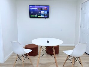 Een kamer met een ronde tafel, twee witte stoelen en een bruin voetenbankje. Een flatscreen televisie is aan de muur boven de tafel bevestigd.
