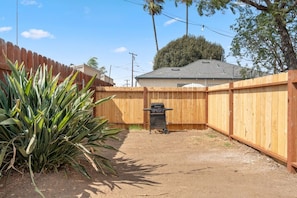 A fenced backyard with a grill, a large plant, and a house in the background.