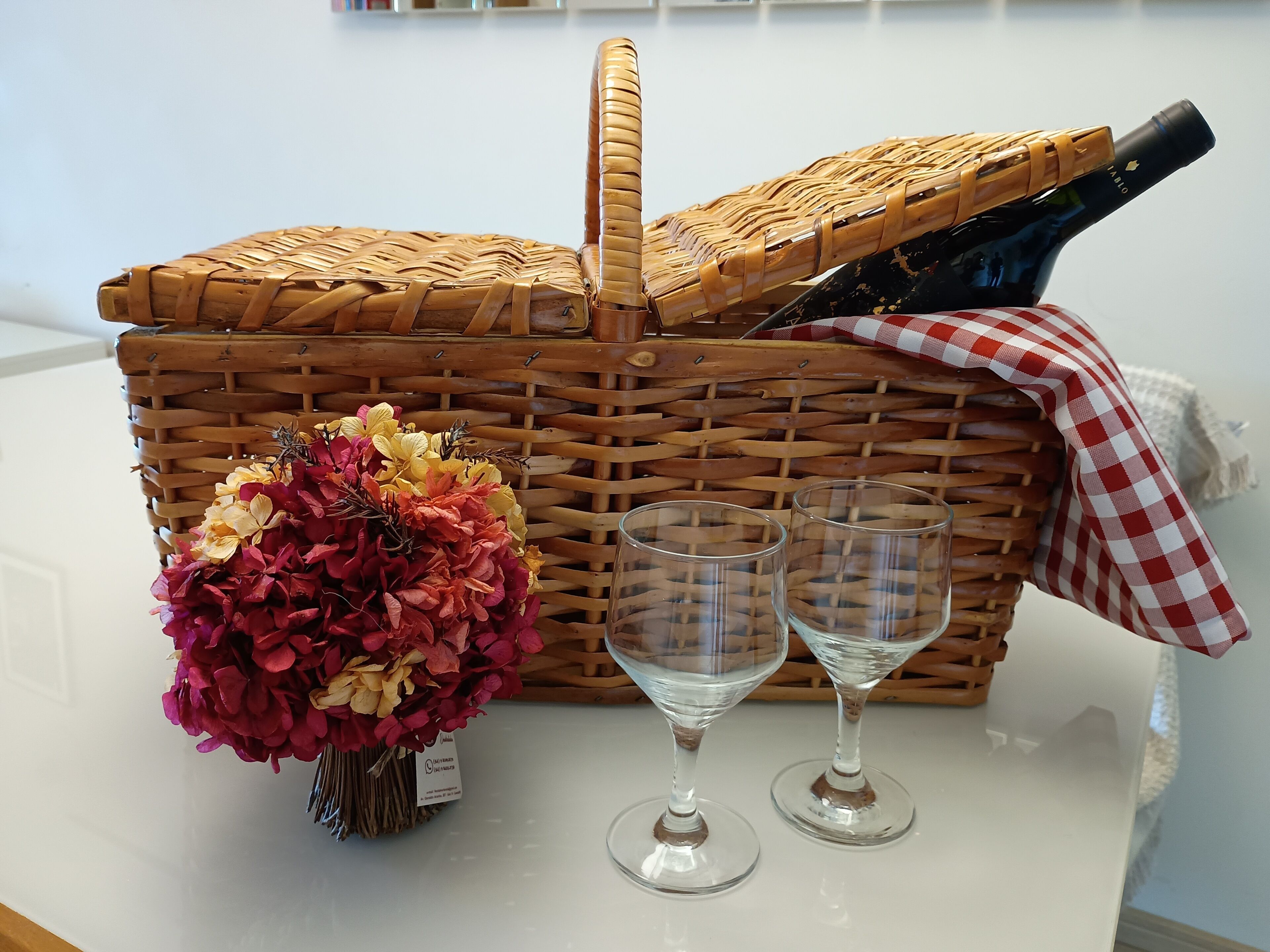 A wicker picnic basket with a bottle and two wine glasses on a white table.