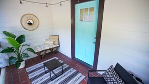 Un porche con una puerta turquesa, un tapiz de pared tejido, un banco con un cojín que dice 'Welcome', una planta en maceta y alfombras estampadas.