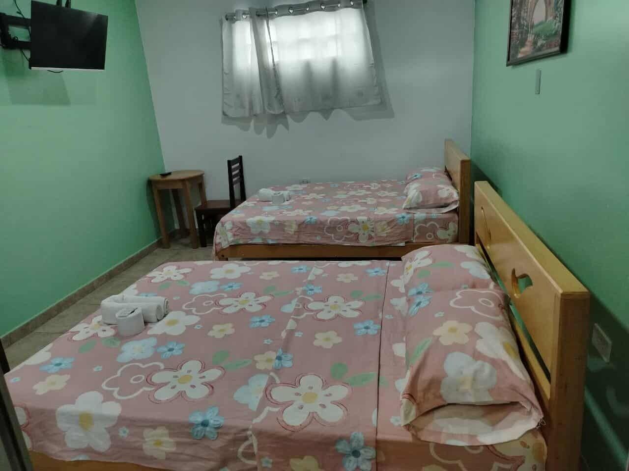 A room with two beds with floral bedding, a small table, a chair, a TV mounted on the wall, and a window with sheer curtains.
