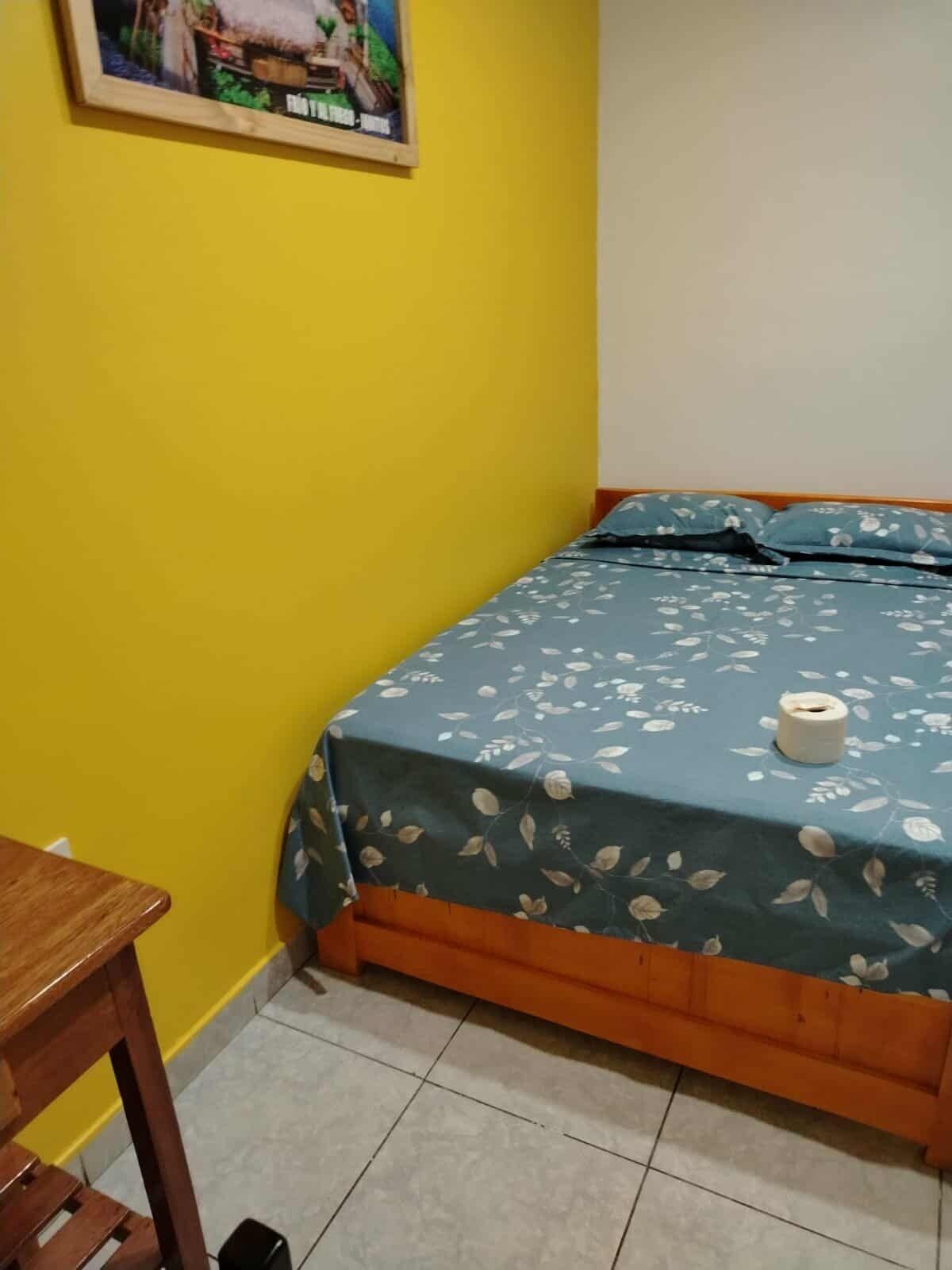 Ein kleiner Raum mit einem Bett, einem Holztisch und einem Bild an der Wand.
