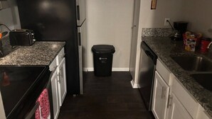 A compact kitchen with a black refrigerator, granite countertops, white cabinets, and a trash bin.