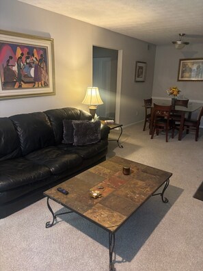 Ein Wohnzimmer mit schwarzem Ledersofa, einem Couchtisch, einer Lampe, einem Essbereich mit Tisch und Stühlen sowie Kunstwerken an den Wänden.