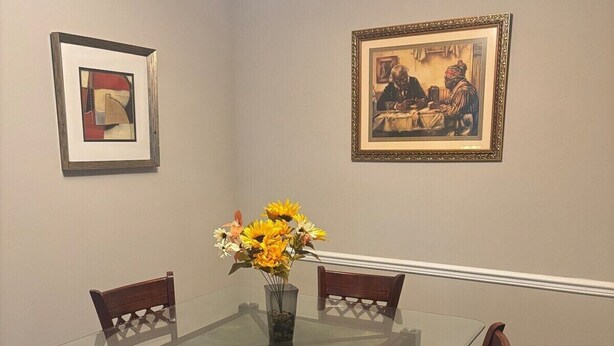 A dining room with a glass-top table, four wooden chairs, and a vase of sunflowers. There are two framed pictures on the walls.