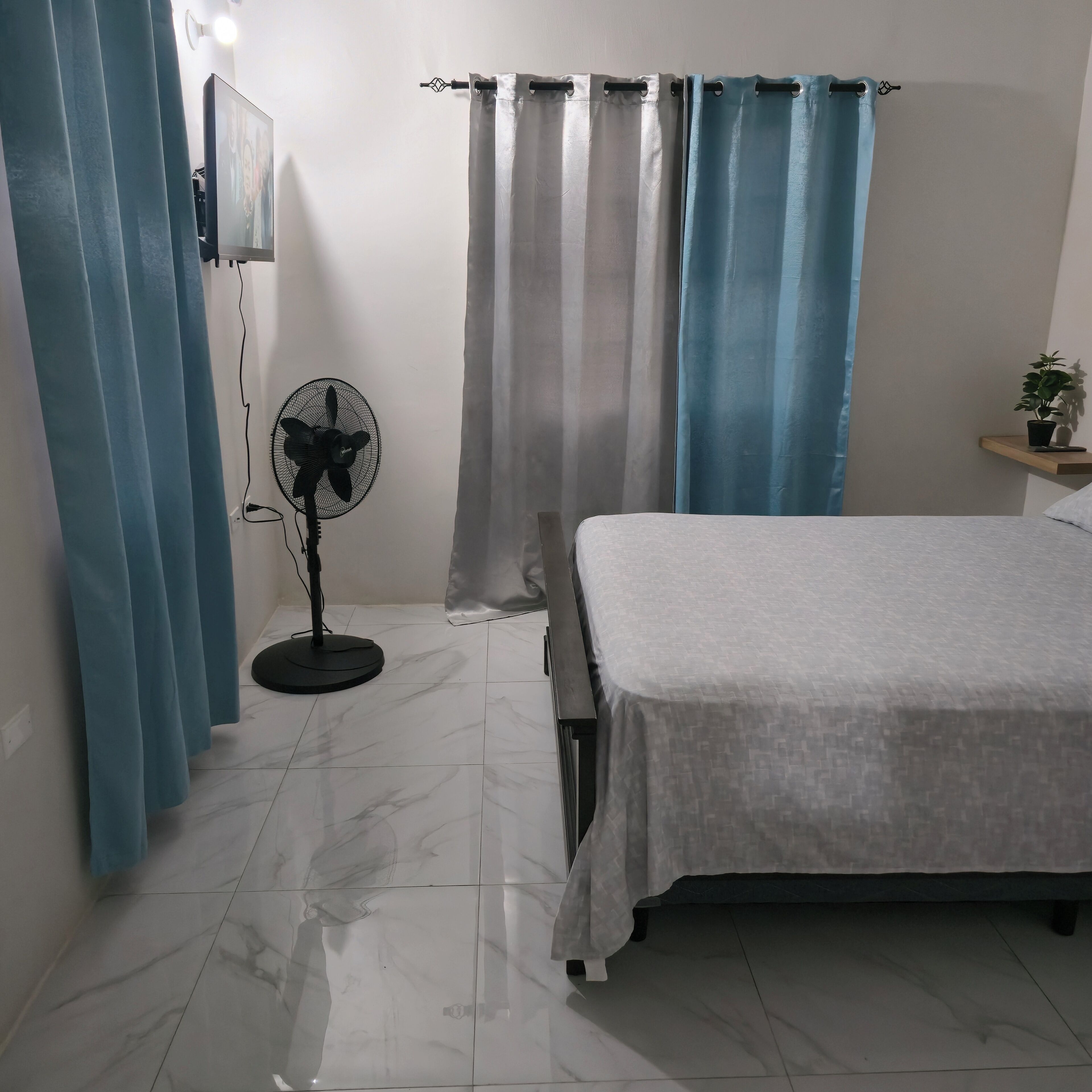 A bedroom with a bed, a standing fan, and curtains.