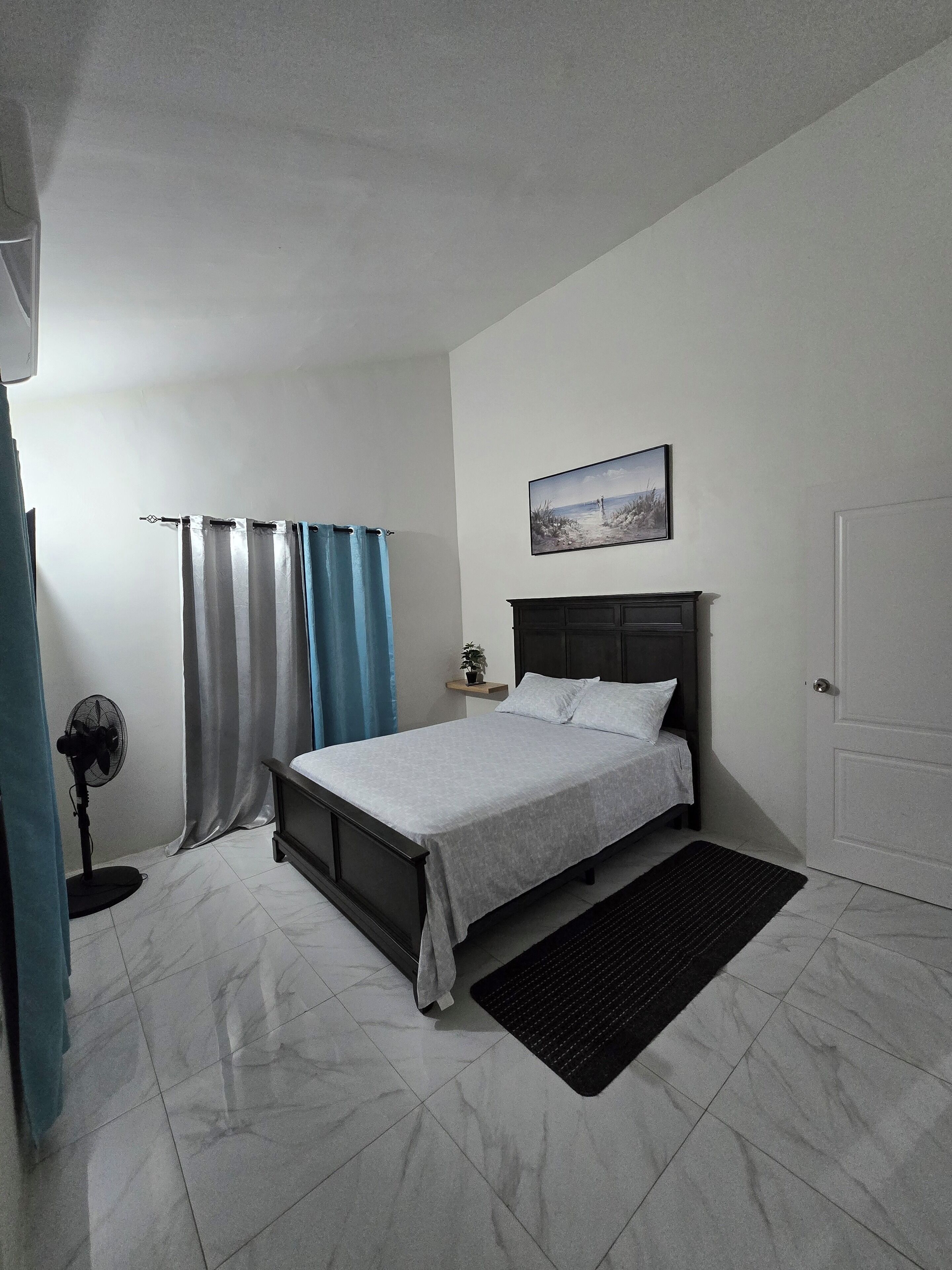 A bedroom with a bed, a bedside table, a wall-mounted picture, a door, and a floor fan.