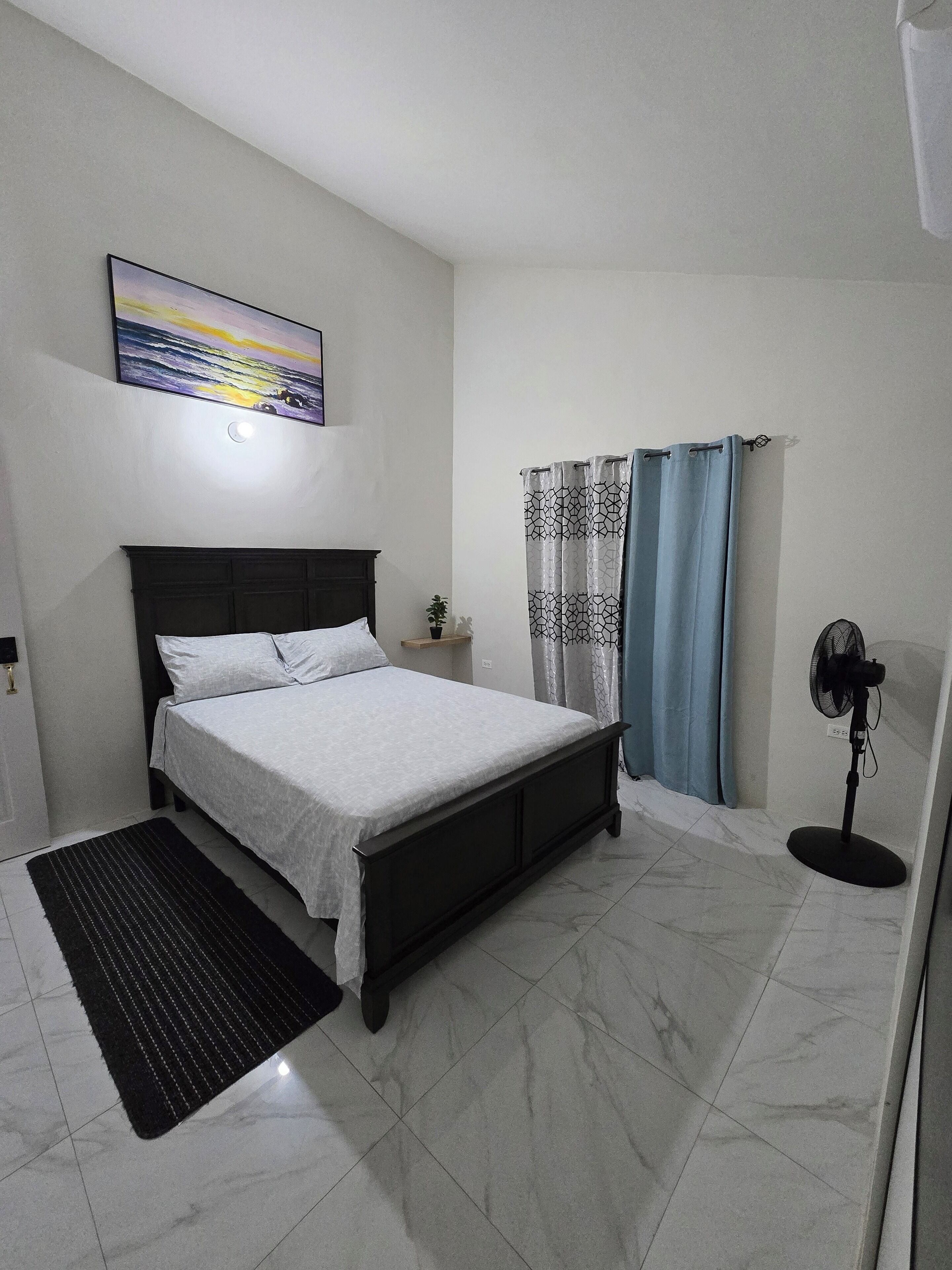 Quarto com cama, mesa de cabeceira, luminária na parede, ventilador de chão e janela com cortinas.