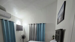 Een slaapkamer met een bed, gordijnen, een televisie, een ventilator, een deur en een kleine plant.