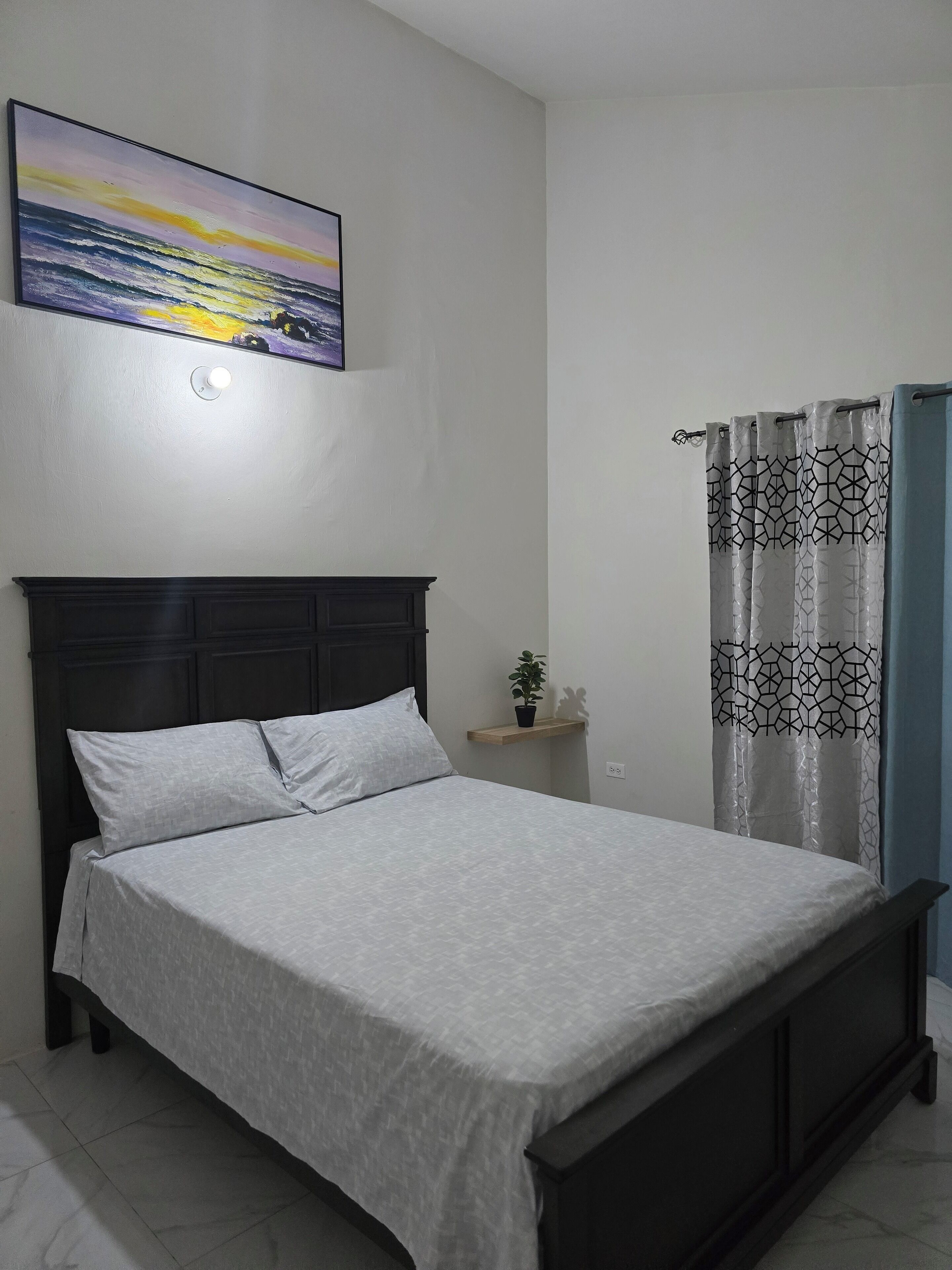 Un dormitorio con una cama, una mesita de noche, una planta en maceta y una ventana con cortinas estampadas.