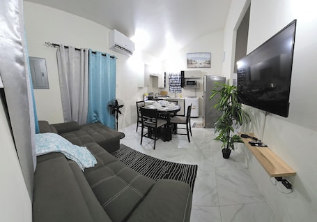 A compact living space with a sofa, dining table, chairs, a TV, a microwave, a refrigerator, and a potted plant.
