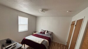 A bedroom with a bed, a desk with a phone and charger, a window with blinds, and a door.