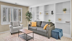 Ein Wohnzimmer mit grauem Sofa, einem kleinen Sessel, einem Couchtisch und einem blauen Hocker. Ein großes Fenster mit Jalousien, ein Kübelpflanze und Regale mit Dekorationen sind ebenfalls zu sehen.