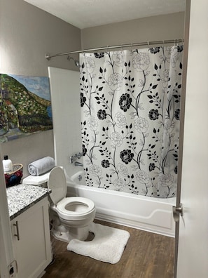 Une salle de bain avec un rideau de douche orné d’un motif floral, des toilettes, un lavabo avec un plan de travail en granit et un petit tableau accroché au mur.