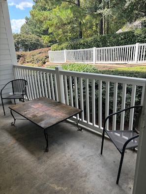 Une terrasse couverte avec une table en métal et deux chaises, entourée d’une clôture blanche et d’une végétation luxuriante.