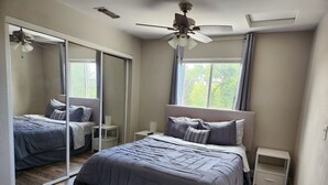 Een slaapkamer met een groot bed, een plafondventilator, een spiegel, een raam met gordijnen en een klein nachtkastje.