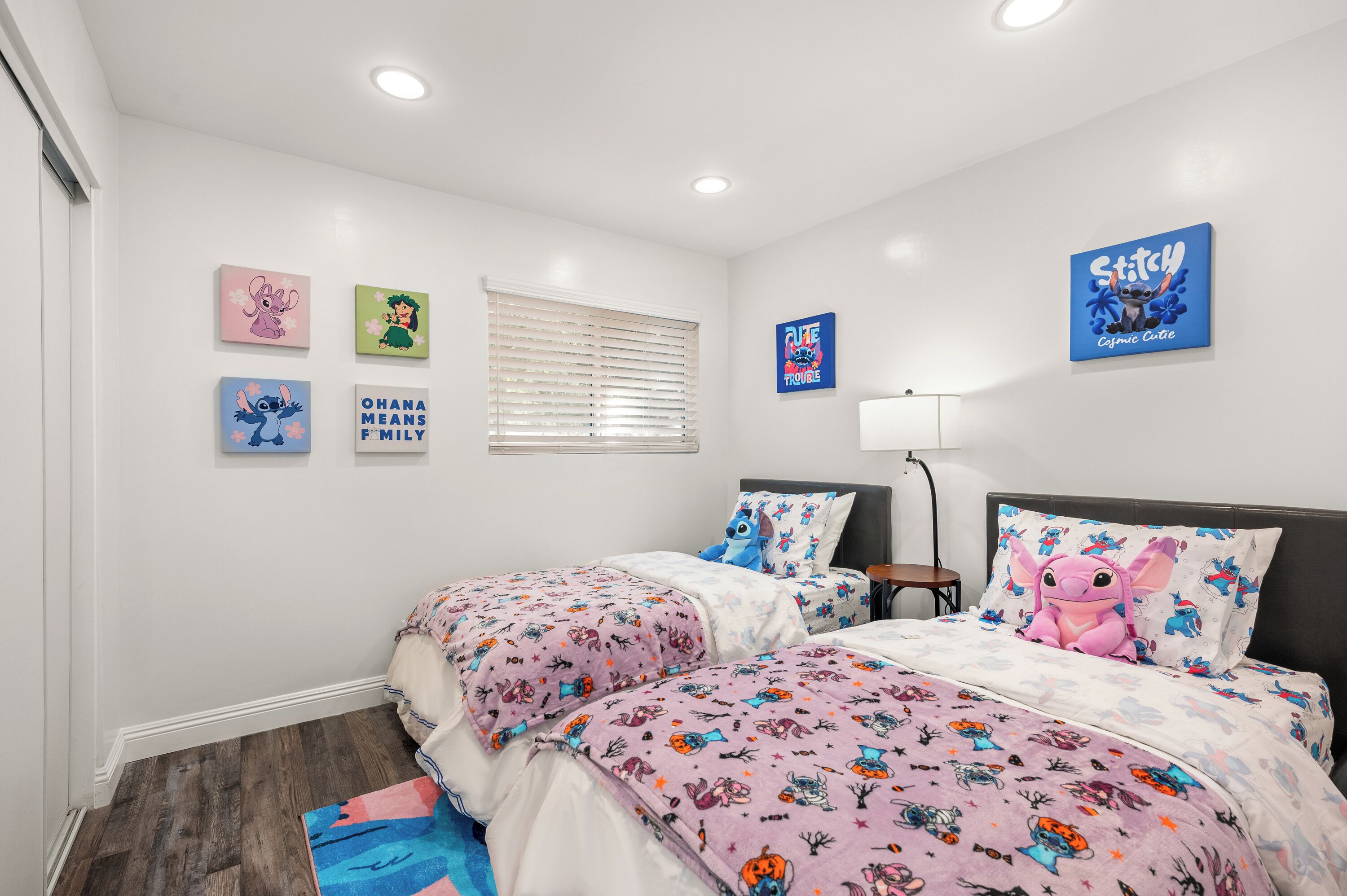 Une chambre avec deux lits, des murs blancs et des luminaires au plafond. On y trouve également des œuvres d’art murales et une fenêtre avec des stores.