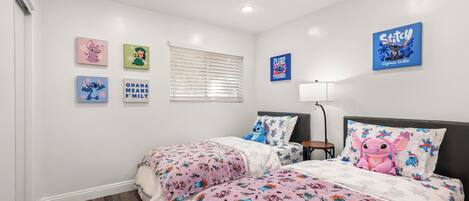 Une chambre avec deux lits, des murs blancs et des luminaires au plafond. On y trouve également des œuvres d’art murales et une fenêtre avec des stores.