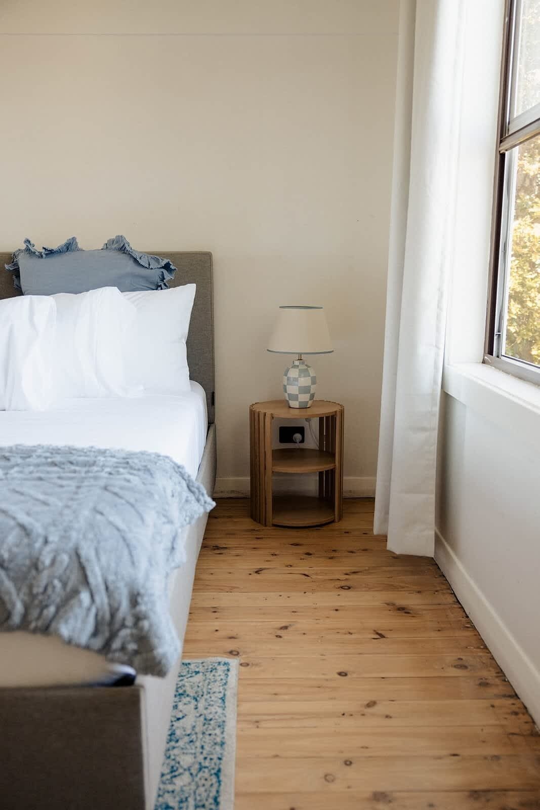 Een slaapkamer met een bed, een nachtkastje met een lamp, een raam en een houten vloer.