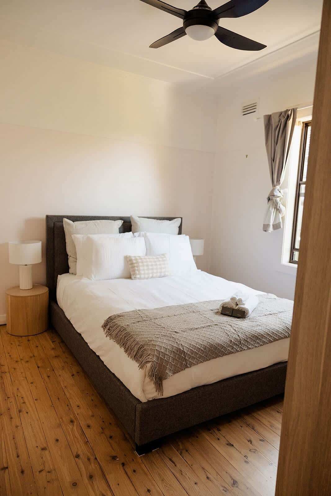 A neatly made bed with white linens and a brown throw, a bedside lamp, and a ceiling fan in a room with wooden flooring and a window with curtains.