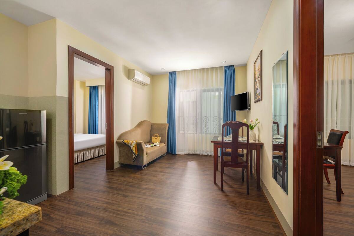 A hotel room with a bed, a sofa, a dining table with chairs, a TV, and an air conditioning unit.