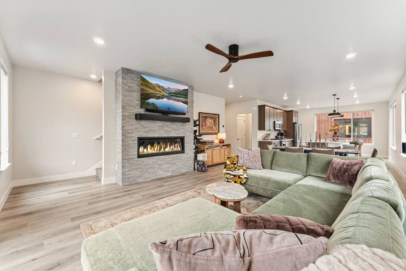 A spacious living room with a fireplace, a large flat-screen TV mounted on a stone wall, a green sectional sofa, and a ceiling fan.