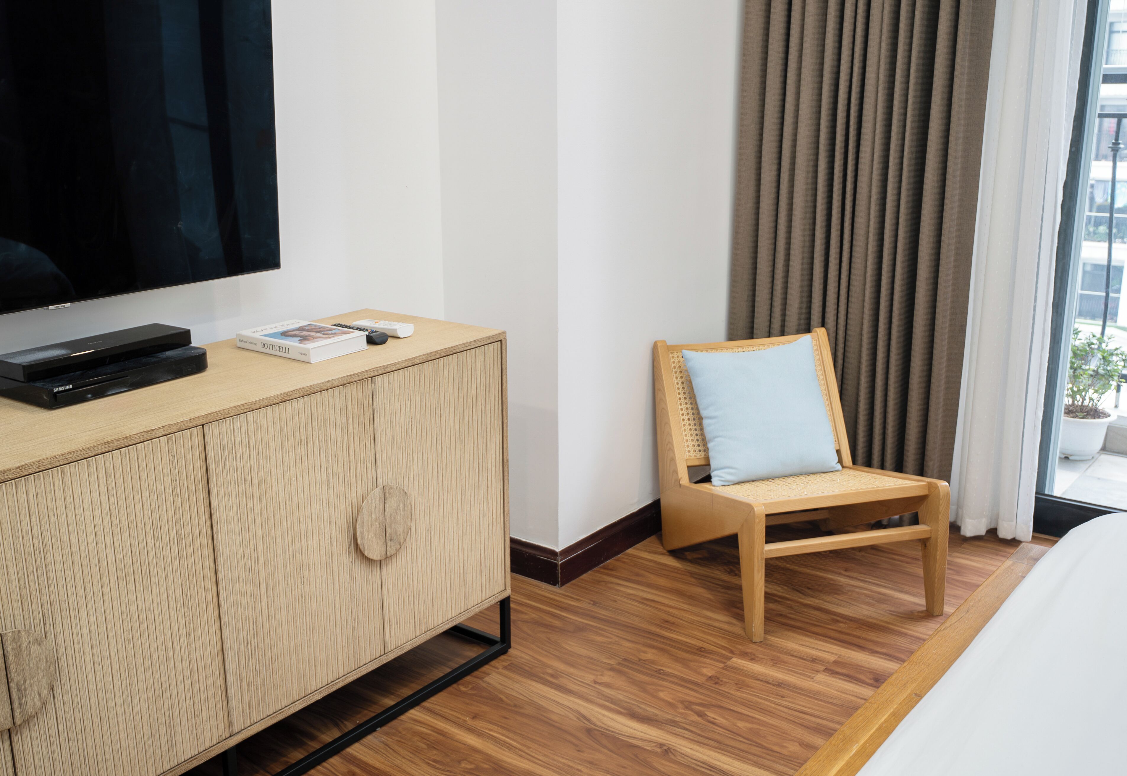A hotel room with a large flat-screen TV, a wooden cabinet with a remote control, a light blue cushioned chair, and a large window with curtains.