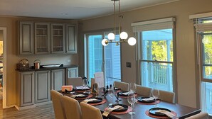 A dining area with a table set for six, surrounded by chairs, in a room with a kitchen in the background and large windows. - SAUNA IS TEMPORARILY OUT OF SERVICE . (Norwich)