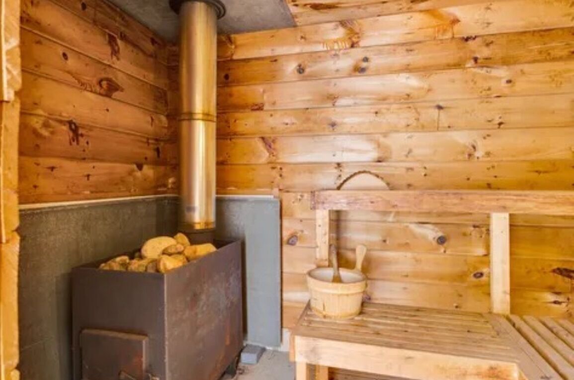 A wooden cabin interior with a wood-burning stove, a metal bucket, and a wooden bench.