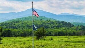 A flagpole with an American flag and a blue flag in a grassy field with a mountain in the background. - White Mountain Cottage, Birchcliff (Shelburne)
