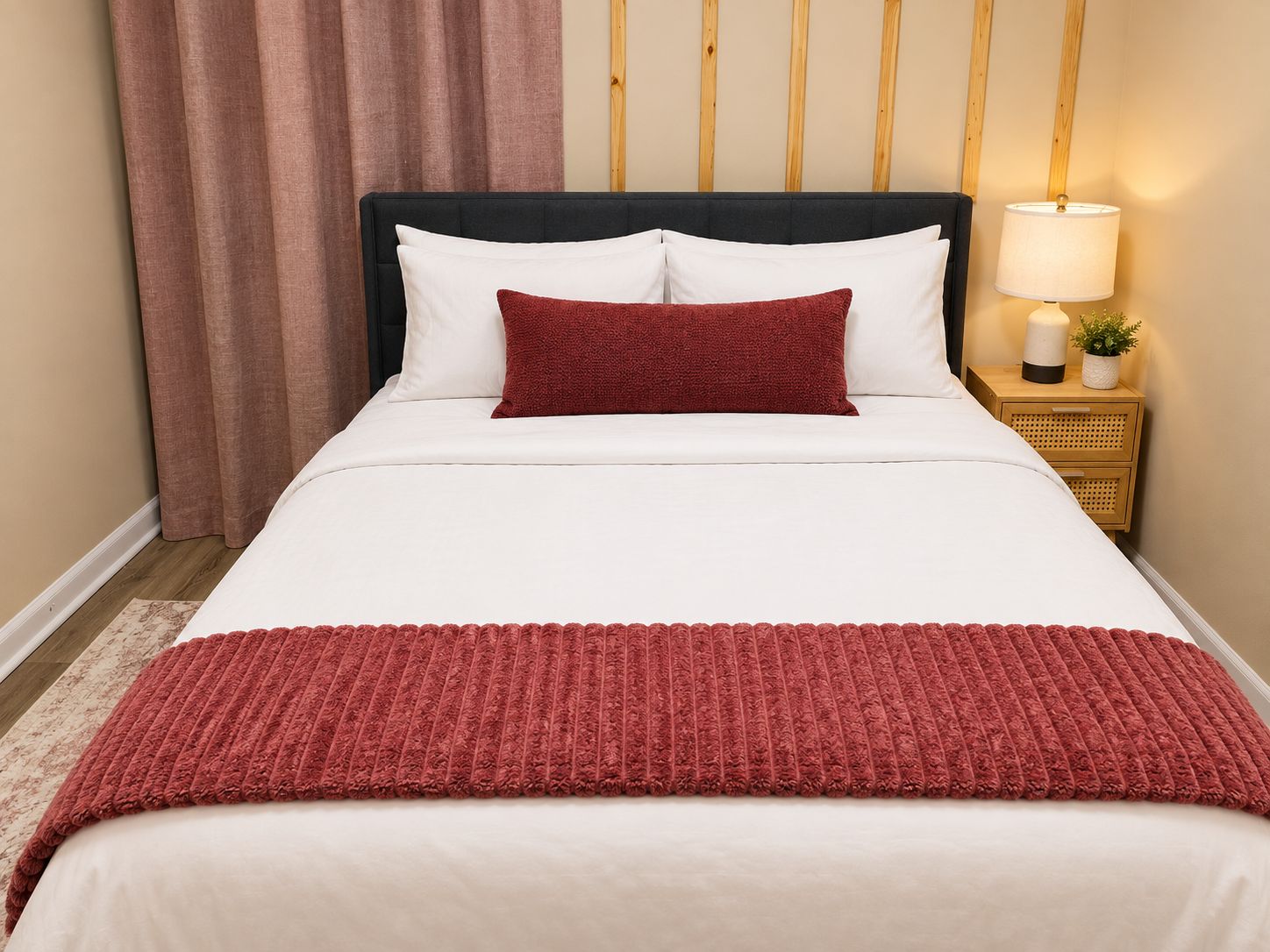 A bed with white bedding and a red throw blanket, a wooden nightstand with a lamp, and a wooden headboard.