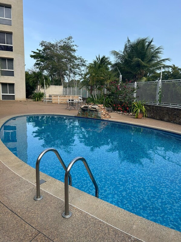 A swimming pool with a tiled deck and metal handrails, surrounded by trees and a stone wall. - Enjoyment at Playa El Angel (Pampatar)