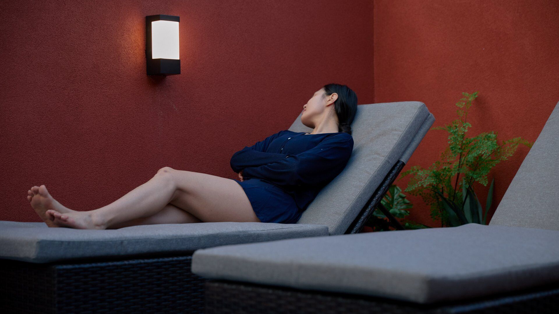 A person lying on a lounge chair in a room with a red wall and a wall-mounted light fixture.