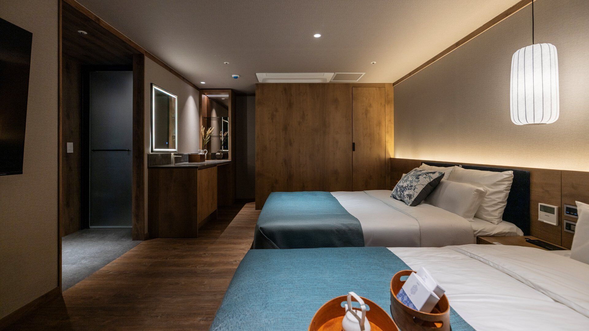 A hotel room with two beds, a wooden headboard, a bedside table, a nightstand, a mirror, and a small kitchenette area.