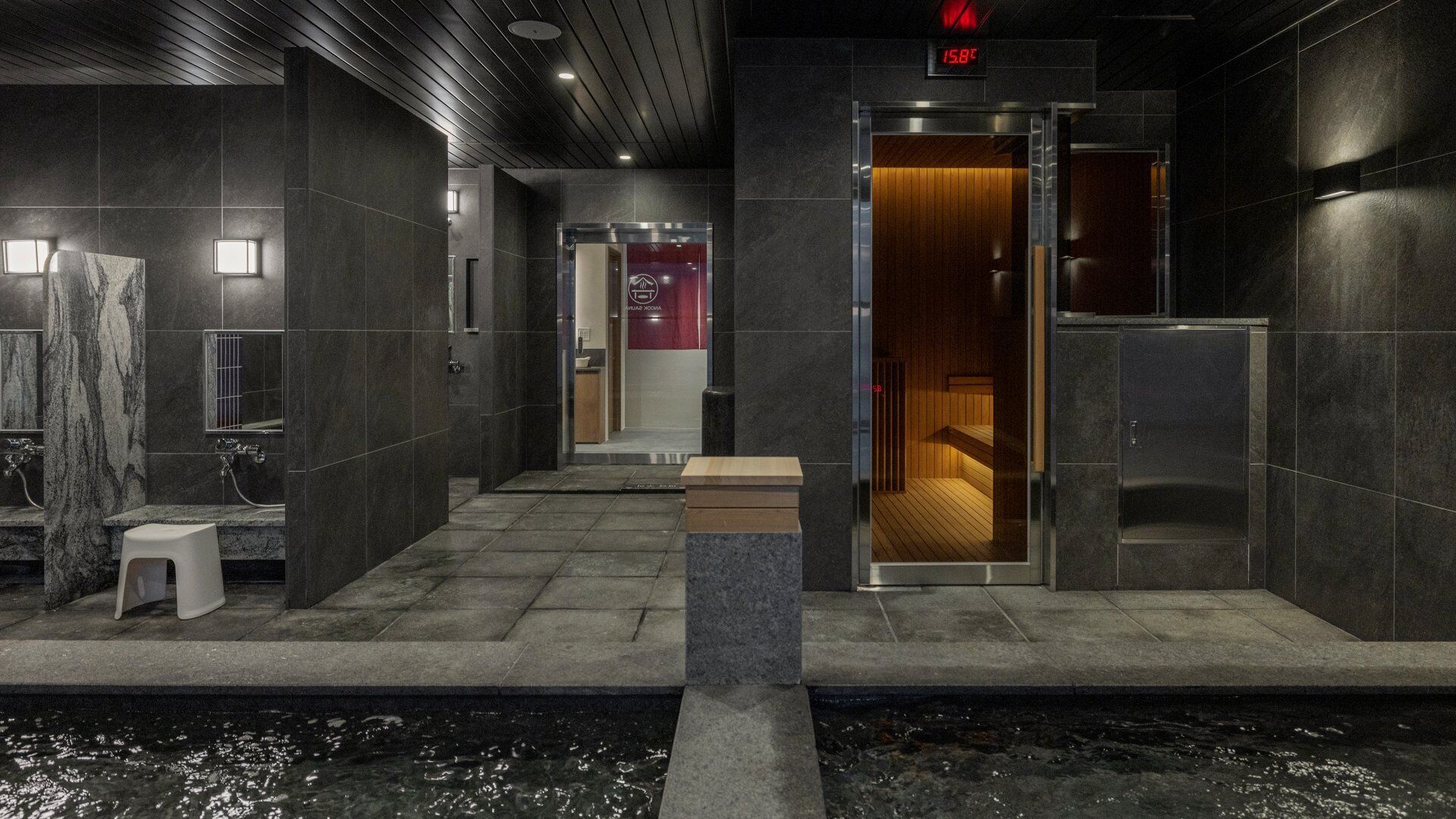 A modern bathroom with dark tiles, a marble sink, a wooden bench, and a sauna room visible through a glass door.