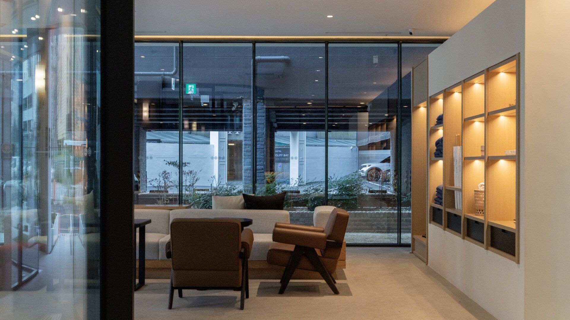 A modern hotel lobby with glass walls, a seating area with sofas and chairs, and a shelving unit with towels.