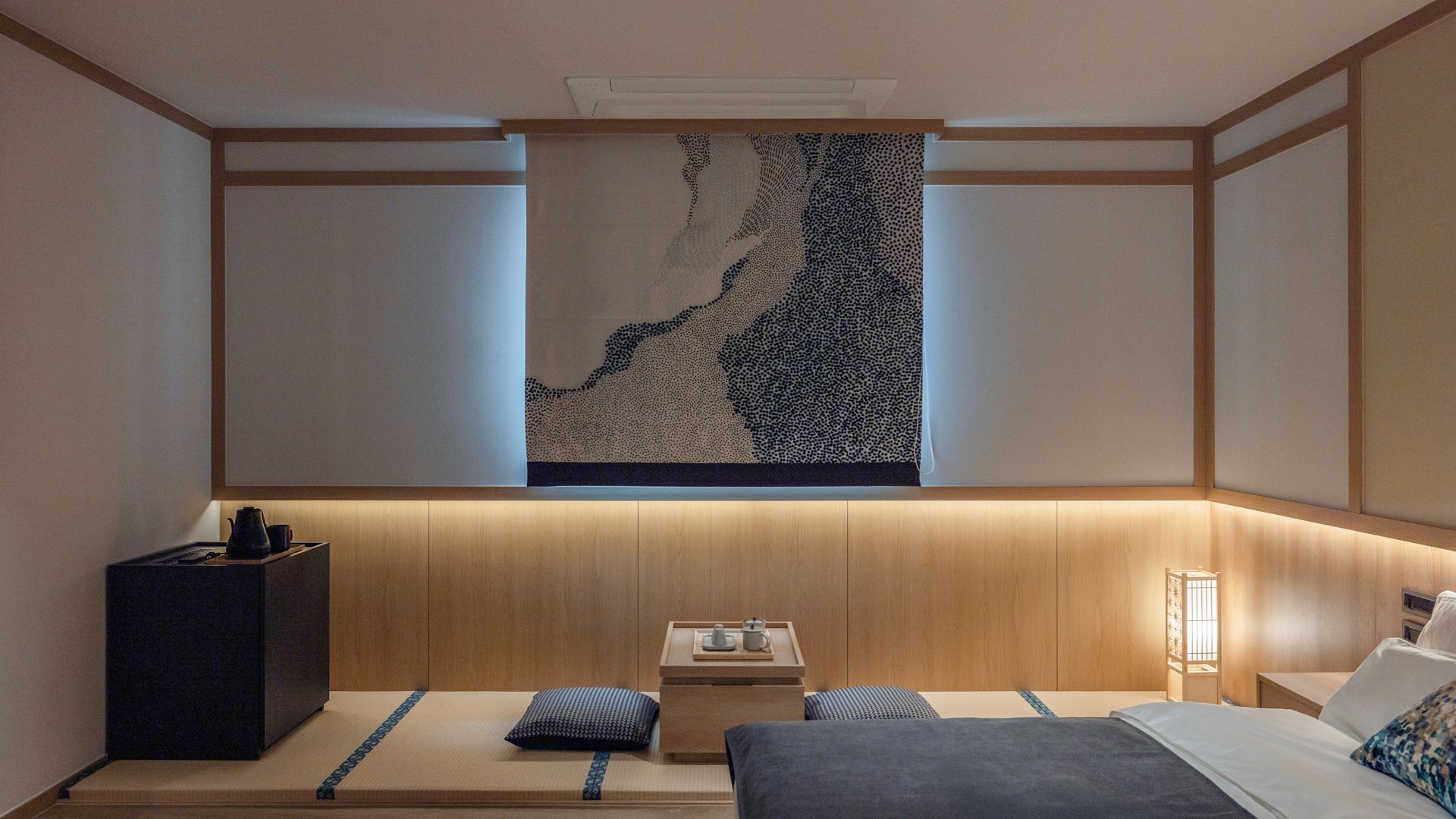 Une pièce de style japonais traditionnel, avec un sol recouvert de tatamis, un lit bas en bois, une petite table avec un plateau et une œuvre d’art fixée au mur représentant un paysage naturel.