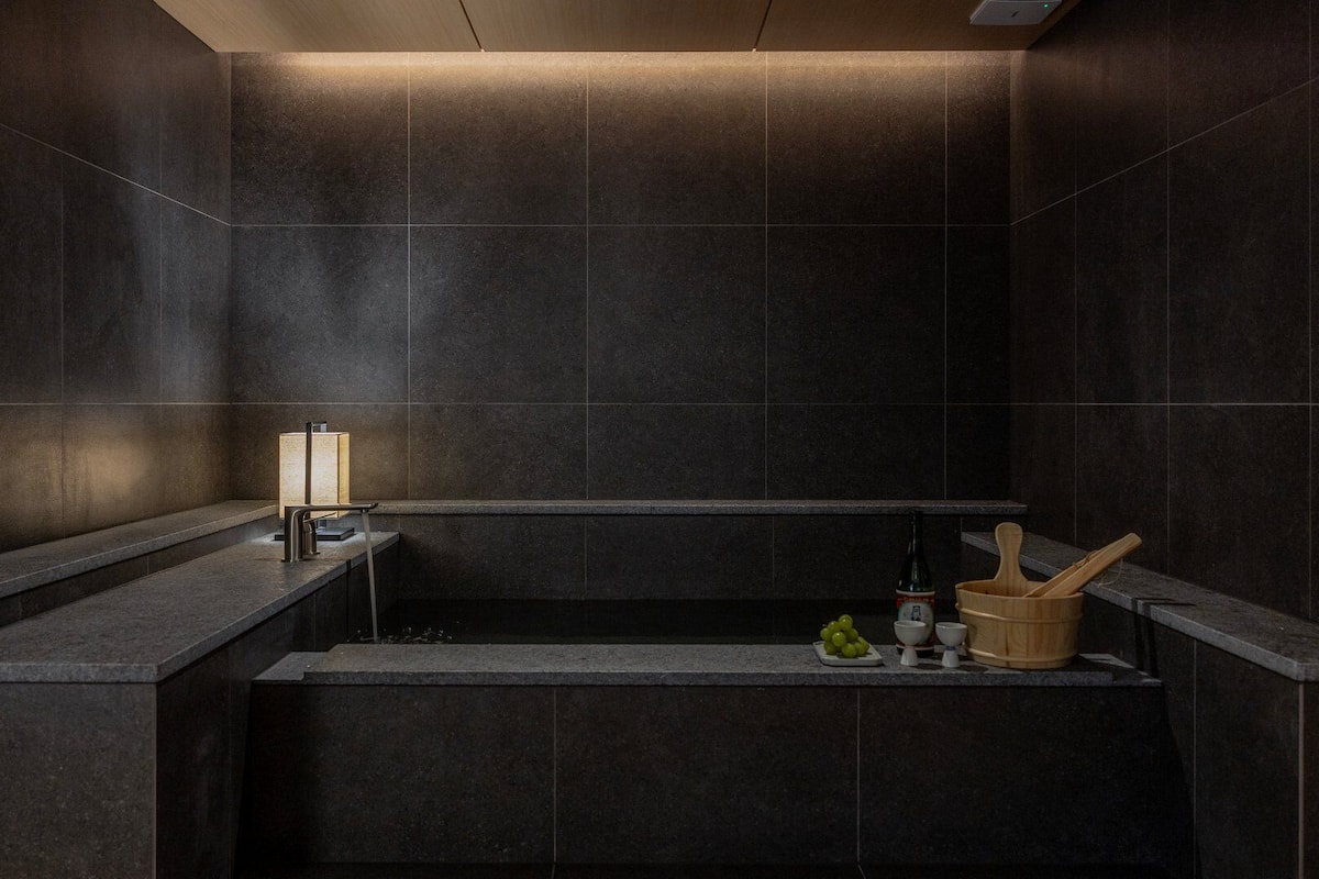 A modern bathroom with dark tiles, a built-in sink, a towel rack, and a wooden bucket.