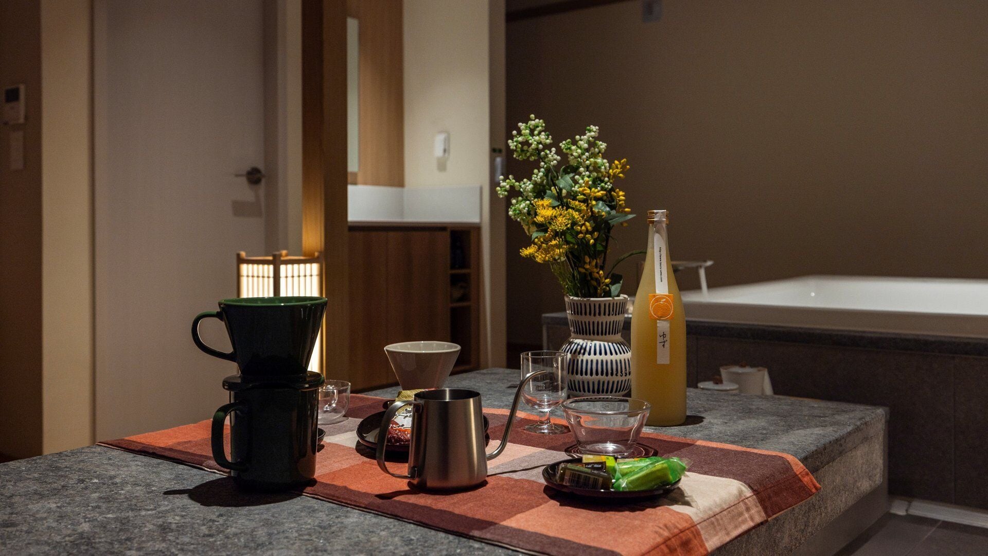 A bathroom vanity with a towel, a bottle of orange juice, a green mug, a glass, and a small plant.