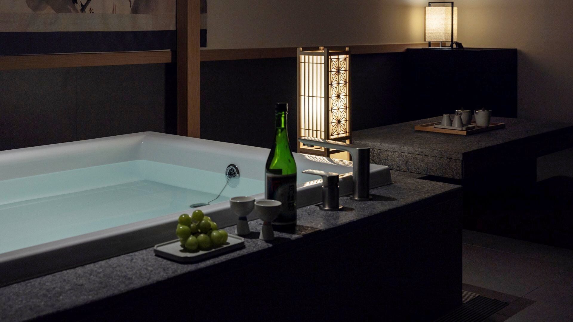 A modern bathroom with a large bathtub, a sink, and a small countertop with a bottle and cups.