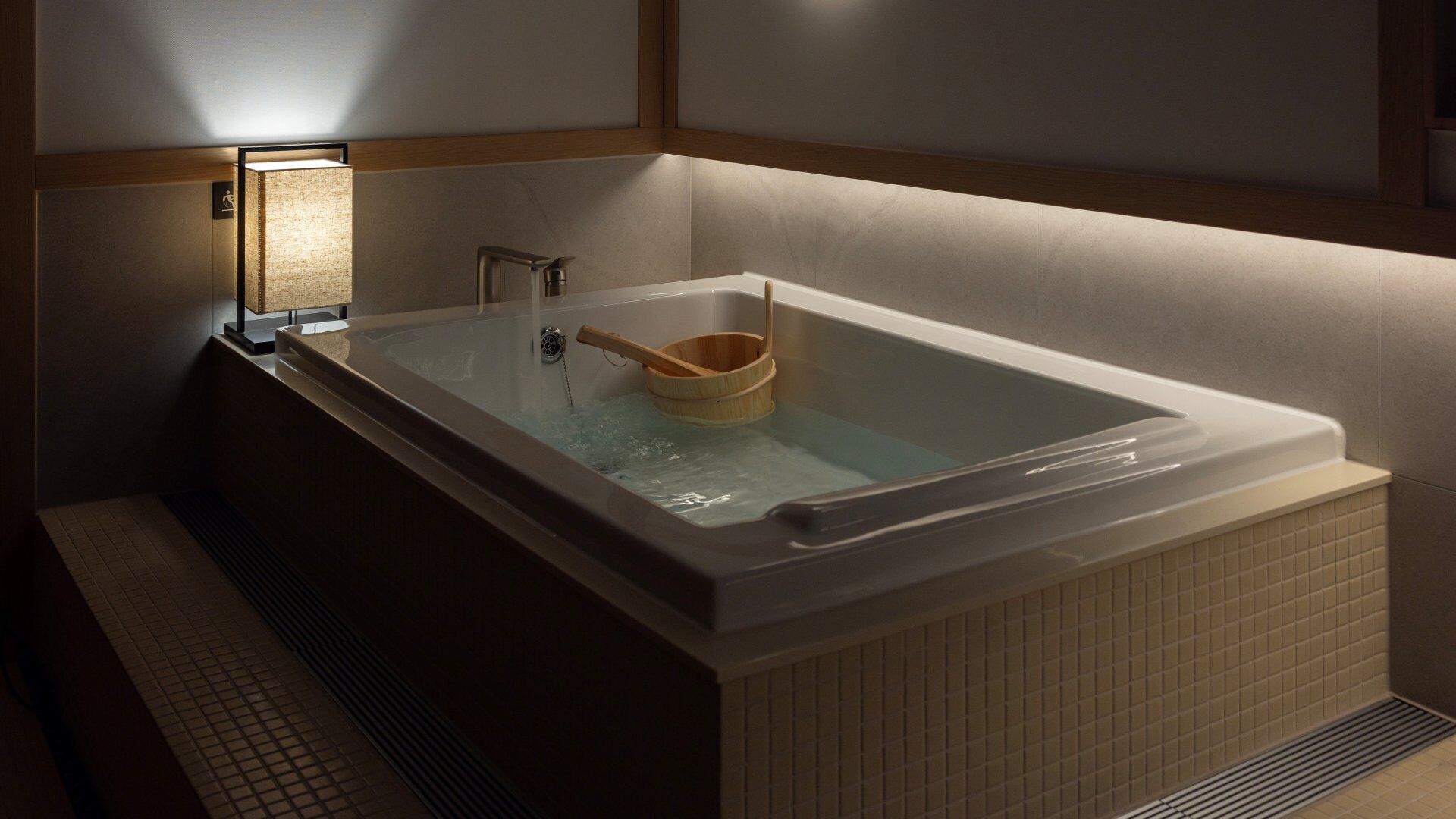 Un baño moderno con una bañera rectangular, un cubo de madera y un aplique de pared.