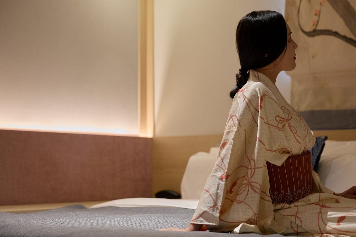 A person in traditional attire sitting on a bed in a room with a plain wall and a painting.
