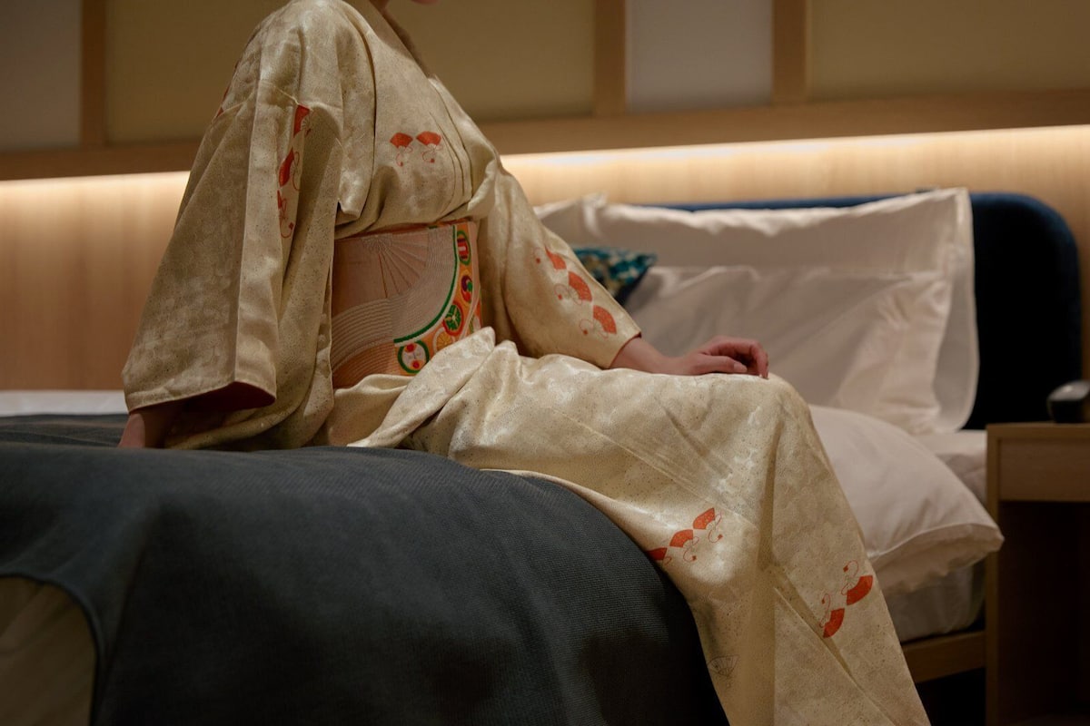 A person in traditional attire sitting on a bed with a wooden headboard, a bedside table with a lamp, and a wall with a patterned design.