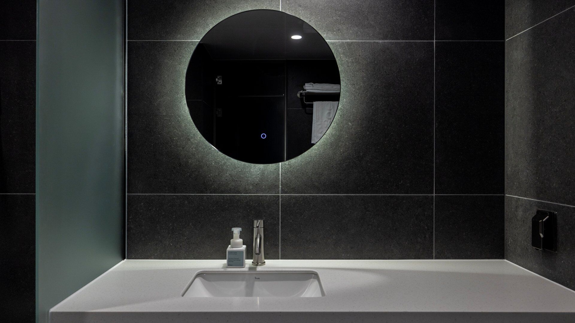 A modern bathroom with a round mirror, dark tiled walls, and a white sink with a faucet.