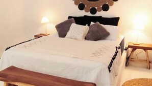 Una cama bien hecha con cubrecama y almohadas blancas, un banco de madera al pie de la cama y dos lámparas de noche sobre mesitas de madera.