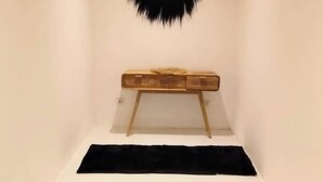 Una habitación estrecha con una pared blanca, un tapete negro, una cesta tejida sobre un soporte de madera y un cojín negro en el suelo.