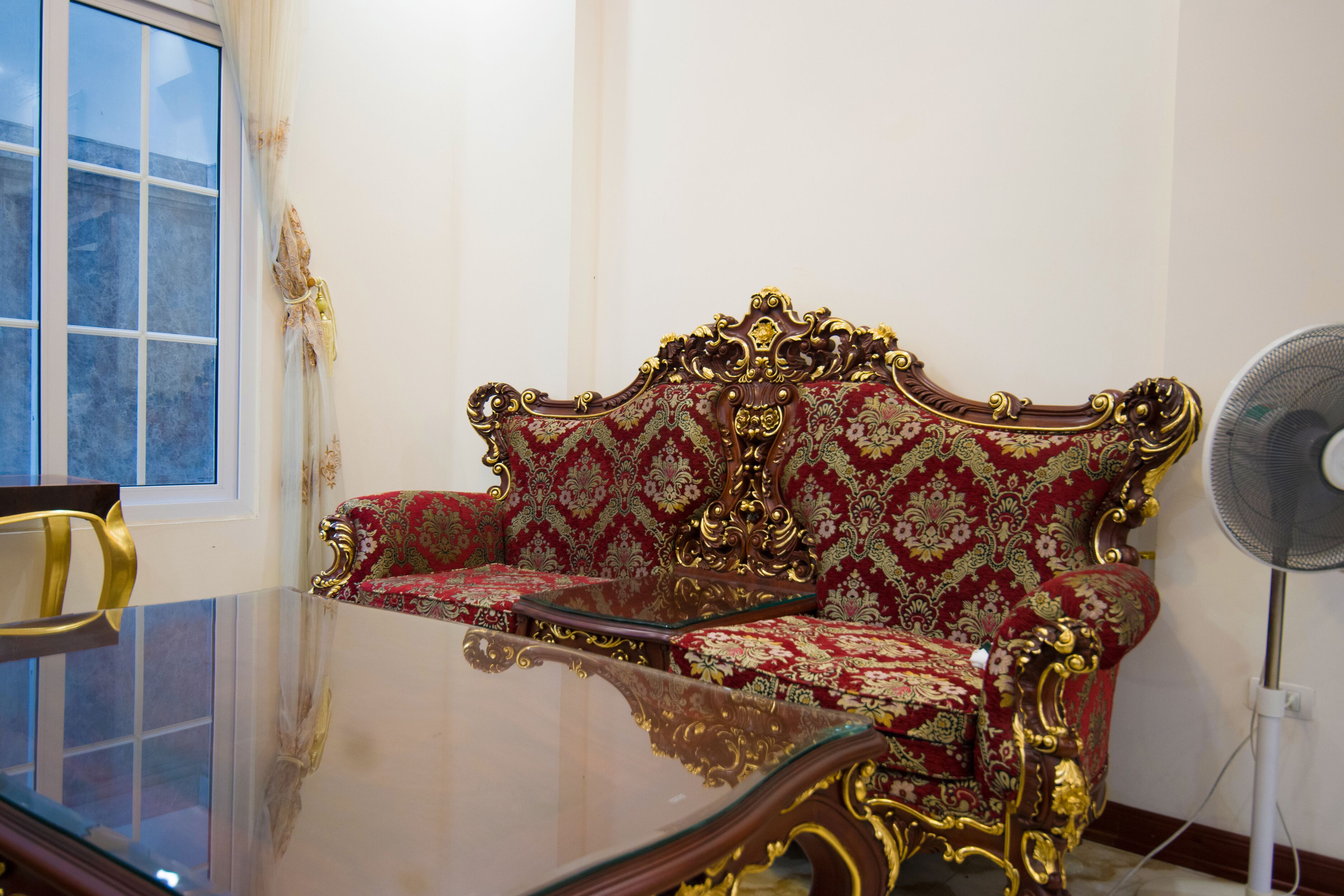 A room with a glass table, a red and gold ornate sofa, a chair, and a standing fan.